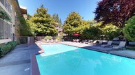 This outdoor amenity space is a standout feature of a well-established multi-story residential community, offering a generous swimming pool as its centerpiece. The pool is framed by classic brick coping and surrounded by a wide, paved sun deck furnished with lounge chairs and shaded seating areas, providing ample space for relaxation and recreation. A separate hot tub is conveniently accessible via a stainless steel handrail, nestled alongside the pool with a dedicated seating area nearby.

The grounds are thoughtfully landscaped with an abundant mix of mature trees, flowering shrubs, rhododendrons, and manicured hedges that create a lush, private atmosphere. Tall deciduous and ornamental trees — including striking red-leafed varieties — surround the perimeter, offering natural shade and a vibrant canopy, particularly during warmer months. The entire pool area is enclosed by a secure metal fence, ensuring safety and privacy.

Adjacent to the pool, a quieter courtyard patio area provides additional outdoor seating set against the building's exterior, surrounded by dense greenery. The multi-story building features private balconies that overlook the pool deck, connecting the interior living spaces to the outdoor environment. Overall, the space exudes a tranquil, resort-like ambiance ideal for outdoor leisure.