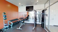 This fitness room is designed to support a well-rounded workout routine within a single, efficiently organized space. The room features a bold orange accent wall that adds energy to the environment, complemented by neutral white ceilings fitted with recessed lighting for bright, even illumination. A large wall-mounted flat-screen television is positioned prominently, allowing users to stay entertained during their workouts.

A full-length mirror along one wall visually expands the room and provides a reflective surface for monitoring form during exercise. The space is equipped with a dumbbell rack holding a comprehensive range of free weights, an adjustable workout bench, and medicine balls for varied training options. A multi-functional cable machine on the opposite side offers additional strength training versatility.

Natural light filters in through a window, enhancing the overall atmosphere. The flooring consists of durable rubber gym matting, providing both safety and comfort. The layout is thoughtfully arranged to maximize usable floor space, making this a practical and functional fitness facility.