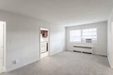 This single-floor residence offers a practical and comfortable layout across several distinct living areas. The living room serves as the heart of the space, featuring neutral-toned carpet, bright white walls, and a large window that allows natural light to fill the room. A radiator unit provides heating, and the open feel of the space allows for flexible furniture arrangements. The separate kitchen is accessible directly from the living area and is equipped with rich dark-espresso cabinetry, granite-style countertops, a gas range, and tile flooring — offering both functionality and a contrasting visual warmth. The bedroom is a generously sized, carpeted room with a ceiling fan light fixture and a closet, providing a quiet and comfortable retreat. A second carpeted room with multiple windows and its own door offers versatility as an additional bedroom or flexible living space, complete with a radiator for heating. The full bathroom features a vanity with a white countertop, wood cabinetry, tile flooring, a tub-shower combination, and a window with natural light. Clean white walls and classic finishes carry throughout the space, creating a cohesive and functional environment.