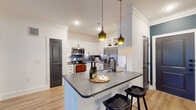 This well-appointed single-floor residence showcases a thoughtfully designed open-concept layout, where the kitchen serves as a central highlight. The kitchen features crisp white shaker cabinetry with matte black hardware, dark granite countertops, a stainless steel appliance suite including a French door refrigerator and range, a dark tile backsplash, and a peninsula with pendant lighting above — creating an inviting and functional cooking space. A hallway connects the main living areas seamlessly, with light-toned hardwood flooring running throughout, lending warmth and visual continuity across the space. Crown molding adds a refined architectural detail to the living areas. The living room is generously proportioned, offering an open canvas for flexible furnishing arrangements. Bedrooms are bright and airy, benefiting from natural light through blinds-fitted windows, ceiling fans, and clean white walls. The bathroom is a standout feature, boasting a granite vanity, matte black fixtures, and a dramatic dark marble-tiled shower and tub surround that brings bold, sophisticated contrast to the otherwise neutral palette. A distinctive dark louvered interior door adds a unique design accent throughout the space.