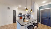 This well-appointed single-floor residence showcases a thoughtfully designed open-concept layout, where the kitchen serves as a central highlight. The kitchen features crisp white shaker cabinetry with matte black hardware, dark granite countertops, a stainless steel appliance suite including a French door refrigerator and range, a dark tile backsplash, and a peninsula with pendant lighting above — creating an inviting and functional cooking space. A hallway connects the main living areas seamlessly, with light-toned hardwood flooring running throughout, lending warmth and visual continuity across the space. Crown molding adds a refined architectural detail to the living areas. The living room is generously proportioned, offering an open canvas for flexible furnishing arrangements. Bedrooms are bright and airy, benefiting from natural light through blinds-fitted windows, ceiling fans, and clean white walls. The bathroom is a standout feature, boasting a granite vanity, matte black fixtures, and a dramatic dark marble-tiled shower and tub surround that brings bold, sophisticated contrast to the otherwise neutral palette. A distinctive dark louvered interior door adds a unique design accent throughout the space.