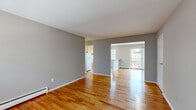 This single-floor unit offers a clean, functional layout with a neutral gray color palette and warm hardwood flooring that runs throughout the main living and bedroom areas. The open living space is generously sized, with natural light filtering in through multiple windows dressed with horizontal blinds. Baseboard heating is present throughout, ensuring year-round comfort.

The kitchen is well-appointed with warm honey-toned wood cabinetry, ample upper and lower storage, a gas range, white refrigerator, and a built-in peninsula with a counter-height surface that connects seamlessly to the living area. The kitchen is finished with tile flooring and receives natural light from a large window, and also features a convenient pass-through shelf. Adjacent to the kitchen, a sliding glass door opens to a private outdoor balcony.

The bedrooms continue the hardwood flooring theme and are finished with the same cool gray tones. Each room is equipped with a window-unit air conditioner and baseboard heaters, offering independent climate control. The primary bedroom includes a walk-in closet and connects to a private hallway leading to the bathroom. The overall space presents a practical and cohesive design, ideal for comfortable everyday living.