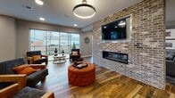 Located at 505 W 7th St in New Castle, Delaware, this single-floor community amenity space combines modern design with inviting, functional areas throughout. The main lounge is anchored by a striking exposed brick accent wall housing a sleek linear fireplace and a large wall-mounted television, creating a warm and social focal point. Richly toned wood-look flooring flows seamlessly throughout the entire space, complementing the contemporary neutral color palette. Large floor-to-ceiling windows and glass doors flood the lounge with natural light and offer views to an outdoor area, enhancing the open and airy atmosphere. Circular pendant lighting adds a bold architectural accent above key gathering zones. A dedicated dining and multipurpose area features curated abstract artwork and connects directly to a fitness center, visible through glass-paned French doors. The game room offers a full-size pool table, a built-in upholstered bench with decorative seating, and wall-mounted game decor, creating an energetic yet comfortable retreat. The kitchenette area stands out with a dramatic veined marble island, dark cabinetry, open wood shelving set against exposed brick, and a full suite of small appliances, making it ideal for community events and gatherings. The overall ambiance balances industrial character with polished, modern finishes.