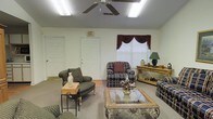 This single-story space features a carpeted common sitting area designed for relaxed gathering and socializing. The room is furnished with multiple plaid-upholstered armchairs and loveseats arranged around a decorative glass-top coffee table with an ornate base, creating a cozy conversational layout. A ceiling fan with integrated lighting anchors the ceiling, complemented by fluorescent light fixtures that ensure consistent, even illumination throughout the room. Natural light filters in through a window dressed with dark burgundy swag curtains and a white blind, adding a touch of traditional elegance to the space. A wooden sideboard along one wall provides decorative display space, holding a table lamp, vase, and small decorative accents. Multiple doorways, including one marked with an illuminated exit sign, suggest the room connects to additional areas of the building. The walls are painted in a neutral off-white tone, maintaining an open and airy feel. The overall ambiance is warm, traditionally styled, and functional, reflecting a comfortable communal environment suited for everyday use.