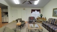 This single-story space features a carpeted common sitting area designed for relaxed gathering and socializing. The room is furnished with multiple plaid-upholstered armchairs and loveseats arranged around a decorative glass-top coffee table with an ornate base, creating a cozy conversational layout. A ceiling fan with integrated lighting anchors the ceiling, complemented by fluorescent light fixtures that ensure consistent, even illumination throughout the room. Natural light filters in through a window dressed with dark burgundy swag curtains and a white blind, adding a touch of traditional elegance to the space. A wooden sideboard along one wall provides decorative display space, holding a table lamp, vase, and small decorative accents. Multiple doorways, including one marked with an illuminated exit sign, suggest the room connects to additional areas of the building. The walls are painted in a neutral off-white tone, maintaining an open and airy feel. The overall ambiance is warm, traditionally styled, and functional, reflecting a comfortable communal environment suited for everyday use.