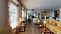 This single-floor clubhouse offers a thoughtfully designed community space that balances comfort and functionality. The main lounge area features rich hardwood-style flooring throughout, complemented by warm, neutral-toned walls that create an inviting atmosphere. Plush seating arrangements, including gold-toned sofas and patterned accent chairs, are arranged around wooden coffee tables, providing an ideal setting for socializing or unwinding. Decorative wall art with nature-inspired motifs and strategically placed floor and table lamps add warmth and character to the space.

A row of large windows along one side of the clubhouse floods the interior with natural light, offering views of the surrounding outdoors. Sheer curtain panels soften the light and contribute to the relaxed ambiance. Lush indoor plants are positioned throughout, enhancing the fresh and welcoming feel of the space.

The clubhouse also features a well-appointed kitchen area with rich dark wood cabinetry, granite countertops, stainless steel appliances, and a peninsula bar with barstool seating — perfect for entertaining or casual gatherings. Recessed lighting throughout ensures the entire space is well-lit. A dedicated corridor connects the various areas seamlessly, reflecting a practical and cohesive layout designed for community use.