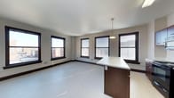 This single-floor space offers an open-concept layout that seamlessly integrates the living and kitchen areas into one cohesive environment. The kitchen features a peninsula with a clean white countertop and dark wood cabinetry below, providing both prep space and a natural divider between the cooking and living zones. A pendant light hangs above the peninsula, adding a focused point of illumination to the area.

The living space is generously proportioned, with light-toned hardwood-style flooring that runs throughout and contributes to the bright, airy atmosphere. Multiple double-hung windows with dark frames line the walls, allowing abundant natural light to fill the interior while offering views of surrounding urban architecture. The windows are fitted with blinds, providing flexible light and privacy control.

The walls are finished in a neutral gray tone, complementing the contrasting dark window trim and baseboards that frame the space with a clean, modern edge. The white ceiling is smooth and uninterrupted, enhancing the sense of openness. Overall, the space presents a practical, contemporary urban layout well-suited for everyday living.