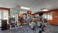 This fitness center is designed to support a comprehensive workout experience within a single, open-plan space. The room is defined by bold terracotta-orange accent walls that lend an energetic atmosphere, complemented by a blue-gray modular carpet floor that adds both comfort and visual contrast. Large wall-spanning mirrors are positioned throughout, enhancing the sense of space and allowing users to monitor their form during exercise.

The cardio section is well-appointed with multiple Precor treadmills, elliptical cross-trainers, and a recumbent stationary bike, all arranged to maximize floor space and usability. For strength training, the facility includes a TuffStuff multi-station cable machine with adjustable attachments, a weight bench, a dumbbell rack, and a stability ball, offering a versatile range of resistance training options. A wall-mounted flat-screen television is available for entertainment during workouts.

Natural light filters into the space through several windows that offer views of surrounding greenery, while flush-mounted panel lighting ensures consistent illumination throughout. Access is provided via both a standard door and a set of double glass exit doors. Sanitation supplies are conveniently wall-mounted near the entrance, and a small waste bin is provided. The overall layout is practical, spacious, and thoughtfully organized to accommodate a variety of fitness routines.