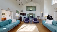 Located at 2828 Marblevista Blvd in Columbus, Ohio, this single-floor community clubhouse is a well-appointed gathering space defined by its dramatic vaulted ceilings and bright, open layout. The main lounge area is anchored by a striking floor-to-ceiling natural stone fireplace, flanked by large windows dressed in soft gold drapes that flood the room with abundant natural light. Hardwood-style flooring runs throughout, complementing the warm, welcoming ambiance. Multiple seating arrangements — including plush teal sofas and deep-toned accent chairs — provide comfortable areas for relaxation and socializing. A mezzanine-level interior window overlooks the main floor, adding architectural depth and visual interest to the soaring interior. The kitchen and bar area features dark cabinetry with a built-in wine rack, a white beadboard bar counter, pendant lighting, and a row of navy blue barstools, making it ideal for casual entertaining. A ceiling fan and recessed lighting enhance comfort throughout the space. The combination of high ceilings, generous natural light, carefully curated furnishings, and distinct functional zones creates a versatile and inviting environment suited for a wide range of community activities.