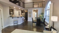 This two-story home showcases an impressive open-concept main level defined by soaring double-height ceilings that create a grand sense of volume and airiness. The living, dining, and kitchen areas flow seamlessly into one another, making the space ideal for both everyday living and entertaining. Dark hardwood flooring runs throughout, providing a rich, warm contrast to the light gray walls.

The kitchen features dark espresso cabinetry, stainless steel appliances, and a granite-topped peninsula with bar seating — a practical and stylish focal point of the main level. The adjacent dining area accommodates a full table and chairs, complemented by a large mirror and decorative greenery that enhance the sense of brightness and depth.

A wrought iron balcony railing on the upper level overlooks the main floor below, adding an architectural elegance while connecting both levels visually. Recessed lighting throughout ensures the space is well-lit, while windows allow natural light to filter in. The overall ambiance is contemporary and polished, combining dark tones with neutral backdrops for a sophisticated aesthetic.