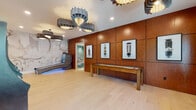 Located at 375 Summer Cypress Cove in Longwood, Florida, this single-floor amenity space is designed for entertainment and leisure, combining contemporary design with a curated selection of recreational features. The open layout is anchored by light-toned hardwood flooring that flows seamlessly throughout, creating a bright and cohesive atmosphere.

A striking feature wall clad in rich, warm-toned paneling serves as a dramatic backdrop for a series of framed fine art prints depicting vintage vacuum tubes, adding an industrial-chic aesthetic to the space. A wooden shuffleboard table is positioned along this wall, offering a classic game option for visitors. Overhead, sculptural layered pendant chandeliers in both blue-grey and dark tones serve as bold focal points, enhancing the room's modern character.

On the opposite end, a large-scale abstract mural in muted tones anchors a lounge area featuring a sleek tanning bed illuminated with ambient blue lighting. A row of upright arcade game cabinets lines one wall, adding a nostalgic and playful element to the entertainment offerings. Floor-to-ceiling windows invite natural light and provide views of the surrounding greenery, while a comfortable seating area nearby offers a relaxed retreat. A clean, well-appointed restroom with tiled flooring and pendant lighting is conveniently accessible within the space.