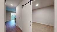 This single-floor space combines contemporary design with practical functionality across its well-appointed rooms. The kitchen serves as a central highlight, outfitted with gray shaker-style cabinetry, a full suite of stainless steel appliances including a refrigerator, electric range, built-in microwave, and dishwasher. A textured tile backsplash adds visual interest, while a movable island with a white countertop provides additional prep and storage flexibility. Wood-look plank flooring runs throughout the kitchen and adjoining areas, lending warmth and cohesion.

A standout architectural detail is the sliding barn door with a matte black hardware track and handle, offering a stylish and space-conscious room divider. The room it accesses is carpeted in a neutral tone, echoing a second carpeted bedroom fitted with a paneled white door featuring black hinges. Both rooms feature neutral wall tones and clean white baseboards, creating a calm, versatile backdrop.

An additional room showcases wood-look flooring and large windows that invite natural light, while a glimpse of a balcony is visible in the background, suggesting outdoor access. Recessed lighting throughout provides consistent, modern illumination. The overall aesthetic is clean, neutral, and move-in ready.