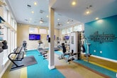 Located in Spring, TX, this fitness center offers a comprehensive and energizing workout environment within a single, open-plan floor. The space is thoughtfully designed with a vibrant, multi-colored striped carpet that adds a dynamic visual appeal, complemented by a bold teal accent wall featuring motivational fitness-themed typography. Recessed lighting and ceiling fans keep the space well-lit and comfortable throughout.

The main workout area is generously equipped with a variety of cardio machines, including treadmills, ellipticals, and stationary bikes, each outfitted with individual screens for entertainment during exercise. A multi-station cable machine and weight bench provide strength training options, while a dedicated free weight rack with a full dumbbell set and adjustable bench caters to a range of fitness routines. Wall-mounted flat-screen televisions are positioned throughout for easy viewing.

Large windows along multiple walls invite abundant natural light and offer views of the outdoor pool area and surrounding greenery, creating an open and airy atmosphere. Crown molding and white structural columns add architectural refinement to the space. A restroom and vanity area with white subway tile, a modern floating sink, and wood-framed glass French doors complete the facility, ensuring both functionality and style.