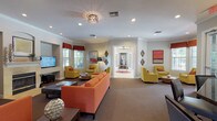 This well-appointed single-floor gathering space is designed to foster comfort and community, offering a cohesive layout with multiple distinct zones that flow seamlessly into one another. The main lounge area is generously proportioned, featuring plush orange sofas and chartreuse accent chairs arranged to encourage conversation and relaxation. Crown molding, recessed lighting, and a decorative flush-mount chandelier lend an air of refined detail throughout the space.

A standout feature is the elegant fireplace, framed by a light stone surround and set beneath an arched alcove, providing a warm focal point in the seating area. A mounted flat-screen television is also integrated into the lounge, adding a functional entertainment element. Large windows dressed with patterned valances allow natural light to fill the rooms while offering views of the surrounding greenery.

An adjoining room, accessible through French doors, provides additional flexible seating space with tile flooring and ample natural light — ideal for meetings or private gatherings. The kitchen is fully equipped with dark espresso cabinetry, granite countertops, stainless steel appliances, and large-format tile flooring, making it well-suited for catering or community events. Throughout, warm neutral wall tones and bold accent artwork create an inviting, energetic atmosphere.