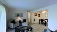 This single-floor residence offers a thoughtfully arranged open-concept layout that seamlessly connects the living and dining areas, creating a cohesive and functional living environment. The living area is generously sized, featuring plush wall-to-wall carpeting that adds warmth underfoot. It is furnished with comfortable seating arrangements centered around a sleek coffee table, complemented by a classic floor lamp that provides ambient lighting. A large framed mirror adorns the wall, enhancing the sense of space and light within the room. Adjacent to the living area, the dining space is defined by a ceiling fan with integrated lighting, offering both comfort and practicality. The dining area accommodates a full-sized table with seating for multiple guests, making it well-suited for everyday meals and entertaining. Framed artwork decorates the walls, adding visual interest and a personalized touch to the space. Neutral-toned walls throughout contribute to a clean, versatile aesthetic that pairs well with a variety of décor styles. A hallway visible from the main living area suggests additional rooms beyond, providing a sense of depth and privacy within the single-floor layout. Overall, the space presents a comfortable and practical living environment with a balanced, inviting character.