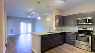 This single-floor space showcases a thoughtfully designed open-concept layout that seamlessly connects the kitchen and living area. The kitchen is a standout feature, equipped with dark espresso-toned cabinetry paired with sleek brushed nickel hardware, offering generous storage throughout. Light granite countertops provide a striking contrast against the darker cabinetry, while a peninsula with an undermount sink and integrated dishwasher adds both functionality and a natural division between the kitchen and living space. Pendant lighting hangs above the peninsula, complementing the track lighting installed along the ceiling for adjustable illumination. A subway tile backsplash adds a clean, contemporary touch to the kitchen walls. The living area extends naturally from the kitchen, bathed in natural light streaming through sliding glass doors that open to an outdoor area. A ceiling fan with a light fixture enhances comfort in this zone. Hardwood-style flooring runs throughout, tying the spaces together cohesively and contributing to the modern, low-maintenance aesthetic of the overall interior.