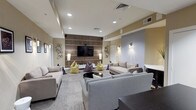 This single-floor recreational space is thoughtfully designed to serve as a social and entertainment hub. The main lounge area is generously proportioned, featuring a large flat-screen television mounted on a striking wood-paneled accent wall with horizontal inlay detailing, creating a strong focal point for the room. Recessed lighting throughout the ceiling provides consistent illumination, complemented by wall-mounted sconce fixtures that add a warm, ambient glow.

Seating throughout the lounge is varied and inviting, combining neutral-toned sofas and loveseats with colorful bean bag chairs in yellow, purple, and gray, encouraging casual, flexible use of the space. Patterned carpet tiles in muted tones cover the floors, adding texture while maintaining a clean, contemporary aesthetic. Decorative geometric wall art and framed prints contribute to the modern character of the space.

A separate gaming alcove connects to the main lounge, identifiable by its "Game Over" wall art and glowing cube accent light. A dark desk and closet storage make this nook functional as well as thematic. A dark console unit provides additional media storage. The overall ambiance is modern, relaxed, and well-suited for group gatherings and entertainment.