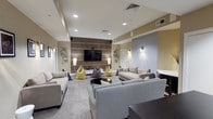 This single-floor recreational space is thoughtfully designed to serve as a social and entertainment hub. The main lounge area is generously proportioned, featuring a large flat-screen television mounted on a striking wood-paneled accent wall with horizontal inlay detailing, creating a strong focal point for the room. Recessed lighting throughout the ceiling provides consistent illumination, complemented by wall-mounted sconce fixtures that add a warm, ambient glow.

Seating throughout the lounge is varied and inviting, combining neutral-toned sofas and loveseats with colorful bean bag chairs in yellow, purple, and gray, encouraging casual, flexible use of the space. Patterned carpet tiles in muted tones cover the floors, adding texture while maintaining a clean, contemporary aesthetic. Decorative geometric wall art and framed prints contribute to the modern character of the space.

A separate gaming alcove connects to the main lounge, identifiable by its "Game Over" wall art and glowing cube accent light. A dark desk and closet storage make this nook functional as well as thematic. A dark console unit provides additional media storage. The overall ambiance is modern, relaxed, and well-suited for group gatherings and entertainment.