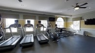 This fitness center offers a thoughtfully designed workout space that balances functionality with refined aesthetics. The room features classic crown molding and a two-tone wall treatment with soft yellow and neutral tones, giving the space an elevated, polished appearance uncommon in typical gym environments.

Natural light streams in through multiple windows, including a distinctive arched window that adds architectural character to the room. Roman shades provide adjustable light control throughout the day. Recessed lighting supplements the natural light to ensure the space remains well-lit during all hours.

The cardio section is equipped with multiple treadmills and an elliptical trainer, each featuring individual display screens for entertainment or workout tracking. A wall-mounted flat-screen television is centrally positioned for shared viewing.

The strength training area includes a well-stocked dumbbell rack with a range of free weights, accompanied by a dedicated weight bench, making it suitable for a full range of resistance training exercises. The dark, rubberized flooring throughout provides durability, safety, and sound absorption, creating a practical workout surface. Overall, the space delivers a complete and comfortable fitness experience within a single, efficiently organized room.