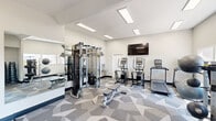Located at 5215 West Peoria Avenue in Glendale, Arizona, this fitness center is a thoughtfully designed, single-floor space that packs a comprehensive workout experience into a compact footprint. The interior features crisp white walls, recessed rectangular lighting panels, and a distinctive geometric-patterned carpet in shades of gray that lends the space a modern, polished aesthetic.

The main workout area is equipped with a Matrix functional trainer cable machine, a flat adjustable workout bench, two elliptical/stair climber machines, and a treadmill — providing a well-rounded selection of both strength and cardio options. A large wall-mounted flat-screen TV is positioned prominently for entertainment during workouts. Full-length mirrors along one wall enhance the sense of space and allow users to monitor their form during exercises.

A dedicated free weights area features a dumbbell rack alongside stability balls and resistance training accessories, catering to a variety of fitness routines. A water bottle filling station is also conveniently located within the workout zone.

An adjacent area transitions to light wood-look flooring and houses a Hub smart package locker system, as well as a wall-mounted mini-split HVAC unit. A glass-panel exterior door in this section provides natural light and direct outdoor access. The overall design is clean, functional, and well-lit throughout.