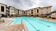 This upscale residential community showcases a compelling blend of Texas Hill Country-inspired architecture, characterized by natural stone facades, board-and-batten siding, and rich red accent detailing across its multi-story buildings. The centerpiece of the property is a generously sized resort-style swimming pool, complete with a tanning ledge, built-in steps, and lane markers, making it suitable for both leisurely relaxation and recreational swimming. An adjacent spa provides an additional retreat within the same courtyard setting.

The expansive pool deck is finished with warm-toned interlocking brick pavers, creating a cohesive and visually appealing outdoor living environment. Comfortable lounge chairs and shade umbrellas are thoughtfully arranged throughout the deck to accommodate residents seeking sun or shade. Meticulously maintained landscaping features tropical palms, ornamental shrubs, and seasonal plantings enclosed by decorative wrought-iron fencing.

A dedicated outdoor lounge area extends from the poolside, offering a covered pavilion equipped with a mounted flat-screen television and an outdoor grill station — ideal for social gatherings. The surrounding multi-story residential buildings form a courtyard configuration, providing both visual enclosure and a sense of community privacy. Overall, this amenity space delivers a well-appointed, resort-caliber outdoor experience.