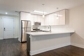 Located at 479 Valley St in Maplewood, NJ, this single-floor unit offers a thoughtfully designed, modern living space with a clean, contemporary aesthetic throughout. The open-concept layout connects the living area and kitchen seamlessly, anchored by rich, dark hardwood flooring that runs throughout the main living spaces. The kitchen is a standout feature, equipped with sleek white upper cabinetry with a high-gloss finish, contrasting wood-toned lower cabinets, and a full suite of stainless steel appliances including a gas range, over-the-range microwave, and refrigerator. A peninsula with a white countertop and an undermount sink doubles as a breakfast bar, illuminated by stylish pendant lighting. The living area is bright and airy, enhanced by a ceiling fan and access to a private balcony through a large sliding glass door that frames views of surrounding greenery. The bathroom is finished with large-format neutral tiles, a soaking tub with a tiled surround, a wood-vanity with white countertop, and recessed lighting, creating a clean and spa-like atmosphere. The overall design balances modern finishes with functional layouts, making this a well-appointed and comfortable space.