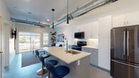 This single-floor urban residence showcases a compelling industrial-chic design that blends raw architectural elements with contemporary finishes. Polished concrete floors run throughout, providing a sleek and cohesive foundation for the entire space.

The open-plan kitchen and living area serves as the heart of the unit. The kitchen is outfitted with two-tone cabinetry — crisp white upper cabinets paired with gray lower cabinets — complemented by a light quartz countertop. A central island with an integrated sink offers seating for multiple guests via modern blue upholstered barstools with metallic bases. Stainless steel and black appliances, including a French-door refrigerator, electric range, and over-the-range microwave, complete the kitchen. Sleek pendant lights hang above the island, while exposed spiral ductwork runs prominently along the ceiling, reinforcing the industrial character. The living area receives natural light from windows that look out to the surrounding neighborhood.

The bedroom features an accent wall in a deep teal-gray tone, an exposed pipe detail along the ceiling, a walk-in closet, and a window with outdoor views, creating a calm and private retreat.

The bathroom includes a wide vanity with white cabinetry and quartz countertop, a full tub-shower combination with white subway tile surround, and access to an adjacent walk-in closet. A bold front entry door in dark navy anchors the entryway with a strong design statement.