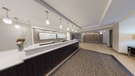 This single-floor lobby presents a polished, contemporary design well-suited for a residential or mixed-use property. The entrance features floor-to-ceiling glass automatic doors branded with the "Avana" logo, offering an immediate view of mature landscaping and a paved exterior forecourt. Upon entry, light-toned tile flooring transitions to patterned carpet, grounding the central circulation area.

The reception desk is a prominent focal point, finished with a dark wood veneer base and a bright white countertop. Above it, a series of cylindrical pendant lights hang at varying heights, complementing two large framed art panels depicting marble-like patterns. Monitors are integrated into the desk for staff use.

To one side, a decorative wall features an arrangement of circular geometric accents in warm gold tones, adding visual texture. Opposite the entry, a seating alcove displays a pair of yellow upholstered benches flanking a rustic wood accent piece, set against sheer floor-to-ceiling curtains that filter natural light from the glazed exterior wall. Tall bamboo plants in dark planters introduce a natural, organic element near the entryway. The overall ambiance is clean, modern, and welcoming, with warm accent lighting enhancing the neutral palette throughout.