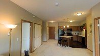 This single-floor space is designed with an open-concept layout that integrates the living and kitchen areas into one cohesive environment. The kitchen is well-appointed with warm honey-toned wood cabinetry, a granite-topped central island with seating for two, and a full suite of black appliances, including a refrigerator, range, and over-the-range microwave. Pendant lighting hangs above the island, adding focused illumination and a subtle decorative touch, while flush-mount ceiling fixtures provide general lighting throughout. The flooring transitions from light-toned carpet in the living area to darker flooring in the kitchen zone, visually distinguishing the two spaces. The walls are finished in a neutral olive-grey tone, complemented by warm wood door trim that adds a cohesive design element. Multiple doorways are visible, with white paneled doors offering access to additional rooms and a darker wood door that serves as an entry point — suggesting a well-connected floor plan. The space maintains a practical, comfortable ambiance suited for everyday living.