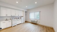 Located at 2274 Newport Blvd in Costa Mesa, California, Unit 305 is a efficiently designed studio that makes thoughtful use of its compact footprint. The main living area features light-toned hardwood-style flooring and bright white walls, creating a clean and airy atmosphere. Recessed lighting illuminates the space, while a window fitted with vertical blinds allows natural light to filter in, enhancing the open feel of the room.

The kitchenette is integrated seamlessly along one wall and is outfitted with white shaker-style cabinetry extending from floor to ceiling, offering generous storage. White appliances including an electric range, microwave, and refrigerator are neatly arranged beneath the cabinetry, complemented by a single-basin sink and ample counter space.

The full bathroom presents a clean, minimalist design with all-white walls and light flooring. It includes a single-vanity with a white cabinet base, a toilet, and a bathtub/shower combination. Stainless steel grab bars are installed near the toilet and along the tub, providing added safety and accessibility. Overhead, a flush ceiling light ensures the bathroom is well lit.

Overall, the unit offers a functional and well-maintained living space with a cohesive, modern aesthetic throughout.