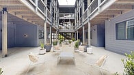 Located at 1115 Seigle Ave in Charlotte's Plaza Midwood neighborhood, this striking space showcases a thoughtfully designed open-air courtyard framed by multi-story industrial architecture. The structure combines steel columns, exposed wood beam ceilings, and metal mesh railings to create a layered, contemporary aesthetic that balances raw industrial elements with inviting outdoor living.

The courtyard serves as the centerpiece of the space, offering generous open-air gathering areas finished with polished concrete flooring. Curated seating arrangements include wire lounge chairs with neutral cushions, low coffee tables, and a long communal dining table, all positioned to encourage both casual relaxation and social interaction. An upper-level balcony with metal stair access overlooks the courtyard, adding vertical dimension and visual interest.

Abundant greenery is integrated throughout via large concrete and ceramic planters filled with a variety of shrubs, grasses, and flowering plants, softening the industrial framework. String lights are strung overhead, contributing warmth and ambiance. Individual unit entries with windows and gray siding line the ground-floor perimeter, while open views to surrounding tree canopies provide a natural backdrop. The overall environment is modern, communal, and well-appointed.