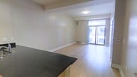 This single-floor space offers a clean, modern layout designed to maximize openness and natural light. The living area features light-toned hardwood flooring that runs throughout, creating a warm and cohesive aesthetic. A prominent floor-to-ceiling sliding glass door floods the space with natural light and provides direct access to an outdoor area, offering views of the surrounding building exterior. White curtains frame the glass door, adding a soft, airy touch to the room.

The kitchen is partially open to the living area and is distinguished by a dark granite countertop that provides a striking contrast against the lighter tones of the space. The cabinetry beneath the counter carries a natural wood finish, complementing the hardwood floors seamlessly.

The interior walls are finished in a neutral off-white tone, contributing to the clean and understated atmosphere throughout. Architectural details include a recessed beam element along the ceiling, adding subtle structural character to the space. Baseboard heating runs along the perimeter, and a wall-mounted climate control unit is also visible, ensuring year-round comfort. The overall ambiance is bright, minimalist, and functional, making effective use of the available floor plan.