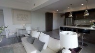Located at 721 N High St in Columbus, Ohio, this single-floor unit sits in the heart of the vibrant Short North Arts District. The space showcases a modern, open-concept design that seamlessly connects the living and kitchen areas, creating a fluid and functional layout ideal for both everyday living and entertaining.

The living area features a clean, neutral color palette with light gray walls and recessed lighting that contributes to the bright, airy atmosphere. A tray ceiling detail adds architectural interest overhead, while the open floor plan allows for generous furniture arrangement, as evidenced by the plush white sofa and coordinating accent pieces.

Transitioning into the kitchen, dark espresso cabinetry provides a striking contrast against the light countertops and stainless steel refrigerator. A peninsula with sleek black bar seating serves as a natural divider between the living and kitchen spaces. A pendant light fixture in a brushed metallic finish hangs above, adding an industrial-modern touch.

A tall, dark wood storage tower anchors the center of the space, offering both practicality and a cohesive design element. Throughout, the neutral tones and contemporary finishes create a polished, sophisticated ambiance.