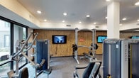 This fitness center offers a thoughtfully designed workout environment suited for a range of exercise routines. The space is anchored by a striking natural-textured accent wall that adds warmth and visual interest to the room, creating a welcoming atmosphere distinct from a typical gym setting. Recessed lighting is evenly distributed across the white ceiling, ensuring the space is bright and well-illuminated throughout.

The facility is outfitted with a robust selection of strength training equipment, including a cable crossover machine, weight bench with a barbell rack, and multiple selectorized resistance machines targeting various muscle groups. Two flat-screen televisions are mounted on the accent wall, providing entertainment and motivation during workouts. The flooring features durable, gray commercial-grade carpet tiles well-suited to a high-traffic fitness environment.

The layout is open and efficient, allowing users to move freely between equipment without feeling cramped. Ventilation is supported by ceiling vents, contributing to a comfortable exercise environment. Overall, this fitness center combines functional equipment with a clean, modern aesthetic, making it a practical and inviting space for daily workouts.