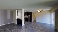 This single-floor space showcases an open-concept layout that integrates the kitchen and living area into one cohesive, functional environment. The kitchen is appointed with dark espresso-toned cabinetry that extends to the upper walls, providing ample storage while creating a bold contrast against the light-colored walls. A stainless steel refrigerator anchors the appliance suite, complementing the modern aesthetic throughout. The kitchen features a peninsula with dark countertops, illuminated by a stylish pendant light that adds a decorative focal point and targeted task lighting. A tile backsplash and an undermount sink complete the kitchen's refined details. The open living area extends generously from the kitchen, unified by rich, dark hardwood-style flooring that runs continuously throughout the space, adding warmth and visual cohesion. A ceiling fan with a light fixture is centrally positioned in the living area, providing both airflow and ambient lighting. Neutral white walls keep the space bright and versatile. A hallway visible from the main area leads to additional rooms, suggesting a practical and well-organized floor plan overall.