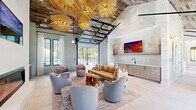 Located at Azalea Landing in Wilmington, NC, this thoughtfully designed community clubhouse offers a generous, open-concept living space that blends coastal sophistication with modern comfort. The single-floor layout is anchored by soaring vaulted ceilings clad in reclaimed wood planking, which lend warmth and texture throughout the main gathering area. A dramatic retractable glass garage door connects the interior seamlessly to the outdoor pool area, flooding the space with natural light and allowing for effortless indoor-outdoor entertaining.

The main lounge area is a standout, featuring a sleek linear electric fireplace framed by a stone surround, built-in cabinetry with open shelving, and a curated arrangement of plush seating. Oversized woven rattan pendant lights hang in a striking cluster above, adding an artistic, coastal-inspired focal point. Large steel-framed windows and glass doors frame lush greenery views, enhancing the airy, nature-connected ambiance.

A dedicated dining area offers a long communal table paired with vibrant green rattan chairs, set against a bold geometric tile accent wall and a wet bar with ample cabinetry. An adjacent lounge nook features softer seating and tall black-framed windows with floor-length draping. Large-format porcelain tile flooring ties the entire space together, creating a cohesive, refined, and inviting atmosphere throughout.