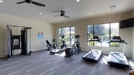 This single-level fitness center offers a clean, functional workout environment designed to support an active lifestyle. The space is outfitted with a selection of cardio equipment, including a treadmill, elliptical trainer, and recumbent bicycle, providing a well-rounded range of exercise options. The flooring features a durable, dark-toned surface suited for fitness use, while the neutral-toned walls contribute to a bright, focused atmosphere. Natural light pours in through multiple large, black-framed windows that look out onto an outdoor pool and patio area, where umbrella-shaded seating is visible among lush greenery — creating a pleasant, resort-like ambiance during workouts. Recessed lighting throughout the ceiling ensures the space remains well-lit at all times of day. Two ceiling fans are installed overhead, promoting air circulation and comfort during exercise sessions. The layout is open and uncluttered, allowing for easy movement between equipment. An informational poster is displayed on the wall, adding a touch of community engagement to the space. Overall, this fitness center combines practicality with an inviting atmosphere, making it a comfortable and motivating place to exercise.