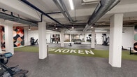 Located at 2500 Ohio Ave in Saint Louis, Missouri, The Koken fitness center is a spacious, single-floor exercise facility offering a comprehensive workout environment. The space features an industrial-inspired aesthetic, with exposed ceiling infrastructure, white-painted concrete block walls, and structural columns that define distinct workout zones throughout the open layout.

A standout feature is the bold, geometric mural anchoring the central wall — a vibrant composition of interlocking circles in orange, black, teal, pink, and green that adds a strong visual identity to the space. A green turf runner extends down the center of the floor, branded with "THE KOKEN" in white lettering, providing a dedicated lane for functional training and movement drills.

The facility is well-appointed with a wide range of equipment. Cardio options include treadmills and a stationary spin bike, accompanied by a wall-mounted television for entertainment. Strength training is supported by a cable/lat pulldown machine, a power tower for bodyweight exercises, a barbell with weight plates, a dumbbell rack, and a selection of medicine balls. Rubber flooring throughout ensures safety and durability across all workout areas.

Overhead lighting illuminates the entire space efficiently, complementing the clean, utilitarian design. The layout provides ample room to move freely between stations, making The Koken a functional and visually engaging fitness destination.
