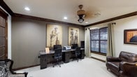 Located in Waxahachie, this single-story space features a well-appointed business center room with a distinctly Western aesthetic. The workspace is anchored by a built-in granite-topped desk that spans the length of one wall, supported by dark-stained cabinetry below providing ample storage. Multiple workstations are accommodated along the counter, each equipped with ergonomic office chairs, making the space practical for shared use.

The room's Western character is established through two large-scale sepia-toned cowboy artworks mounted prominently on the neutral gray walls, complemented by rich dark wood crown molding that frames the space with traditional elegance. Recessed lighting provides even illumination throughout, while a decorative wood-toned ceiling fan with a lantern-style fixture adds both function and rustic charm overhead.

Natural light filters in through a sizable window dressed with horizontal blinds and neutral linen curtains, offering light control and privacy. Soft carpeting in a light neutral tone covers the floor, contributing to a comfortable and quiet working environment. Overall, the space balances professional functionality with a warm, regionally inspired character.