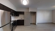 This single-floor unit offers an efficient, open-concept layout that seamlessly integrates the kitchen and living area into one cohesive space. The kitchen is outfitted with dark espresso cabinetry that extends along two walls, providing ample upper and lower storage. Light-toned countertops contrast the dark cabinets, creating a balanced, modern aesthetic. Stainless steel appliances, including a range and microwave, add a contemporary touch, while a single-basin sink is neatly positioned beneath the cabinetry along the back wall.

The flooring throughout is light-toned laminate wood, contributing to a clean and modern feel. Neutral grey-white walls keep the space feeling open and bright, complemented by flush-mount ceiling lighting that ensures even illumination. A partial wall separates the kitchen from the adjoining living area, where an open closet with a hanging rod offers convenient built-in storage. A paneled interior door in a warm greige tone adds a subtle design element and provides access to the closet space. The overall ambiance is clean, functional, and move-in ready, suited to practical, everyday living.