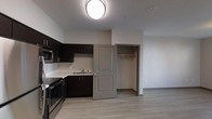 This single-floor unit offers an efficient, open-concept layout that seamlessly integrates the kitchen and living area into one cohesive space. The kitchen is outfitted with dark espresso cabinetry that extends along two walls, providing ample upper and lower storage. Light-toned countertops contrast the dark cabinets, creating a balanced, modern aesthetic. Stainless steel appliances, including a range and microwave, add a contemporary touch, while a single-basin sink is neatly positioned beneath the cabinetry along the back wall.

The flooring throughout is light-toned laminate wood, contributing to a clean and modern feel. Neutral grey-white walls keep the space feeling open and bright, complemented by flush-mount ceiling lighting that ensures even illumination. A partial wall separates the kitchen from the adjoining living area, where an open closet with a hanging rod offers convenient built-in storage. A paneled interior door in a warm greige tone adds a subtle design element and provides access to the closet space. The overall ambiance is clean, functional, and move-in ready, suited to practical, everyday living.
