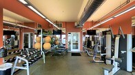 This fitness center is designed to support a comprehensive workout experience within a single, open-plan floor. The space is immediately distinguished by its bold terracotta-red accent walls, which create an energizing atmosphere complemented by a neutral ceiling with exposed ductwork — a nod to industrial-style design that adds both character and a sense of vertical openness. Overhead fluorescent strip lighting ensures the entire area is brightly and evenly illuminated throughout the day. Floor-to-ceiling mirrors line the walls, visually expanding the space and allowing users to monitor their form during workouts. The facility is well-stocked with a wide selection of dumbbells neatly arranged on dedicated racks, a variety of stability and medicine balls, adjustable workout benches, and multiple resistance and weight machines positioned along the perimeter. Rubber flooring throughout provides cushioning, durability, and safety for high-impact activities. Mounted flat-screen televisions are strategically placed for viewing convenience during cardio or strength training sessions. A glass-paneled entry door allows natural light to filter into the space, adding a welcoming touch. Overall, the layout is thoughtfully organized to maximize both functionality and ease of movement.