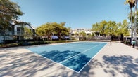 This outdoor recreational space centers around a full-sized tennis court finished in a striking two-tone blue surface with crisp white court lines and a regulation net. The court is enclosed by a chain-link fence, providing both safety and an open, airy feel. Towering palm trees and mature deciduous trees line the perimeter, casting dappled shade across the surrounding walkways and lending the space a tropical, resort-like ambiance. The lush greenery creates a natural buffer that enhances privacy while contributing to a tranquil atmosphere. Set within a well-established residential community, the court is framed by multi-story buildings featuring classic architectural detailing, including covered balconies and decorative awnings, visible in the background. The surrounding landscaping is neatly maintained, with manicured shrubs and ground cover bordering the court's perimeter path. The open sky above allows for abundant natural light throughout the day, making this an inviting venue for both casual play and more competitive matches. The overall setting combines functional sport infrastructure with a carefully curated outdoor environment, offering residents an attractive and accessible amenity.