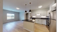 Located at 12191 Felch St in Holland, Michigan, this single-floor unit offers a thoughtfully designed layout with modern finishes throughout. The heart of the space is an open-concept kitchen and living area, where light hardwood flooring extends seamlessly across the room, creating a bright and cohesive atmosphere. The kitchen is well-appointed with crisp white cabinetry, dark countertops, a full suite of stainless steel appliances including a refrigerator, range, microwave, and dishwasher, and an undermount sink. A central kitchen island with a dark countertop and beadboard paneling serves as a functional focal point, illuminated by a pair of stylish pendant lights overhead. The open living area adjacent to the kitchen offers generous space and natural light from a side window. The bedroom features soft carpeting and includes access to a walk-in closet with built-in shelving, providing practical storage. The bathroom is finished with a light wood vanity, white countertop, large mirror, tub surround, and durable tile-style flooring. Overall, the unit presents a clean, contemporary aesthetic with consistent attention to detail across every room.