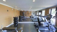 This fitness room is thoughtfully designed to support a well-rounded exercise routine. The space features warm, golden-toned walls complemented by crown molding and recessed lighting, lending the room a polished, finished appearance. A full-length mirrored wall runs along one side, visually expanding the space while allowing users to monitor their form during workouts. The dark rubberized flooring provides a durable, slip-resistant surface suited for a variety of exercises. On the free weights side, a tiered dumbbell rack holds an organized selection of weights, accompanied by a flat bench and a multi-function cable machine, offering strength training versatility. The cardio section is equipped with a treadmill, an elliptical trainer, and a recumbent-style machine, catering to varying fitness preferences and intensity levels. Natural light filters in through windows along the far wall, creating a bright and energizing atmosphere. The combination of practical equipment layout, reflective surfaces, and thoughtful design elements makes this fitness room a functional and inviting space for daily exercise.