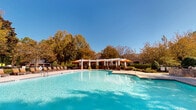 Located at Cameron Woods in Apex, North Carolina, this outdoor amenity area is centered around a generously sized, freeform swimming pool with crystal-clear turquoise water. The pool is flanked by a wide, paver-stone deck that accommodates a generous arrangement of lounge chairs with orange cushions, ideal for sunbathing and relaxation. A white pergola structure provides a shaded gathering area complete with patio tables and vibrant orange umbrellas, offering a comfortable retreat from the sun. A separate poolside building with a brick and siding exterior serves as a clubhouse or amenity structure, providing additional facilities for residents. The pool area is enclosed by a sleek black iron fence that ensures both security and an unobstructed view of the surrounding mature trees, which display rich autumn foliage of gold and amber. A charming gazebo is also visible beyond the fencing, nestled among neatly manicured shrubs and landscaping that enhance the natural ambiance. The space is thoughtfully designed to blend functional outdoor recreation with an inviting, resort-style atmosphere, set against the backdrop of a clear blue sky and the natural canopy of North Carolina's mature hardwood trees.