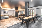 Located at 1669 Scott Blvd in Decatur, Georgia, this single-floor amenity space is thoughtfully designed to serve as a social and recreational hub. The interior showcases a contemporary aesthetic defined by dark-tiled structural columns, a charcoal ceiling, and herringbone-patterned flooring that transitions seamlessly across the various activity zones.

The game area is a central highlight, equipped with a full-size pool table and a shuffleboard table, both positioned beneath stylish pendant lighting clusters that add warmth to the darker design palette. Surrounding the game zone, layered area rugs define comfortable lounge seating areas furnished with teal velvet sofas and accent chairs, creating inviting spots for relaxed gatherings.

A built-in bar area with dark brick tile, wooden accents, and red bar stools anchors one end of the space, complemented by wall-mounted televisions. A Scrabble-inspired decorative board and patterned feature wall add personality to the opposite lounge area.

Floor-to-ceiling glass doors and large windows flood the space with natural light while offering direct views of a landscaped courtyard. The overall ambiance balances energetic entertainment with relaxed social comfort, making it a versatile and well-appointed communal destination.