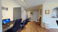 This single-floor space is thoughtfully arranged to support both collaborative and focused work environments. The layout flows between two primary areas connected by hardwood-style flooring that runs throughout, lending warmth and visual continuity to the space.

The workstation area is outfitted with a long, built-in desk along the wall, accommodating multiple ergonomic office chairs and individual workstations divided by low privacy panels. Track lighting runs along the ceiling, providing focused, adjustable illumination across each workspace. A corkboard for notices and memos is mounted on the wall, and the area includes a separate interior door providing access to additional rooms or utilities.

Adjacent to the workstation zone is a spacious conference and gathering room anchored by a large, dark-wood dining-style table surrounded by upholstered chairs. A wall-mounted flat-screen television serves as a focal point, while a ceiling fan with integrated lighting contributes to comfort and airflow. Natural light enters through a broad set of windows, brightening the room and offering views of the surrounding exterior. A decorative arched pass-through opens to a small kitchenette or service counter area, adding convenience for longer meetings or gatherings. Framed artwork adorns the walls, giving the space a professional yet welcoming ambiance throughout.