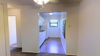This single-floor space offers a straightforward and functional layout designed for comfortable everyday living. The living area is outfitted with a ceiling fan and light fixture, providing both airflow and ambient lighting. An open archway connects the living space directly to the kitchen, creating an accessible and open feel between the two areas. The kitchen features white cabinetry lining one wall, offering ample storage space, along with a sink, a refrigerator, and additional appliances including what appears to be a stove or oven unit on the opposing wall. A window at the far end of the kitchen allows natural light to filter in, brightening the space. The kitchen is finished with hardwood-style flooring that extends throughout, providing a clean and cohesive look, while the living area transitions to a carpeted surface. The walls throughout are painted in a neutral white tone, lending the space a bright and clean aesthetic. The overall design is practical and efficient, making good use of the available layout to deliver a well-connected living and cooking environment.