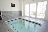 This enclosed spa room offers a dedicated relaxation amenity centered around a generously sized indoor whirlpool. The hot tub is finished with crisp white tile and accented by a decorative blue diamond mosaic border, giving it a clean, polished appearance. A stainless steel entry handrail provides safe and convenient access into the water.

The room features vaulted ceilings that lend a sense of openness to the space, while the lower half of the walls is clad in neutral gray square tile, providing a durable and cohesive aesthetic. The upper walls are painted white, keeping the interior bright and airy. Recessed lighting is installed overhead to supplement natural light.

Large multi-pane windows line one side of the room, allowing ample natural light to fill the space while offering views of an outdoor pool and landscaped surroundings beyond a perimeter fence. A dedicated entry door provides direct access to and from the facility.

Spa rules are posted on the wall, indicating this is a shared amenity space. Maintenance equipment is stored on-site, reflecting active upkeep of the facility. The light-toned tile flooring throughout ties together the overall clean and functional design of the room.